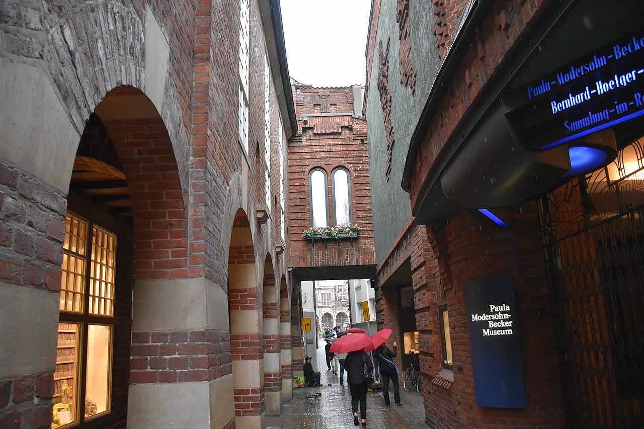 The narrow red brick Böttcherstraße in the rain, with arches on the left, a brick bridge between the buildings and people with red umbrellas walking away