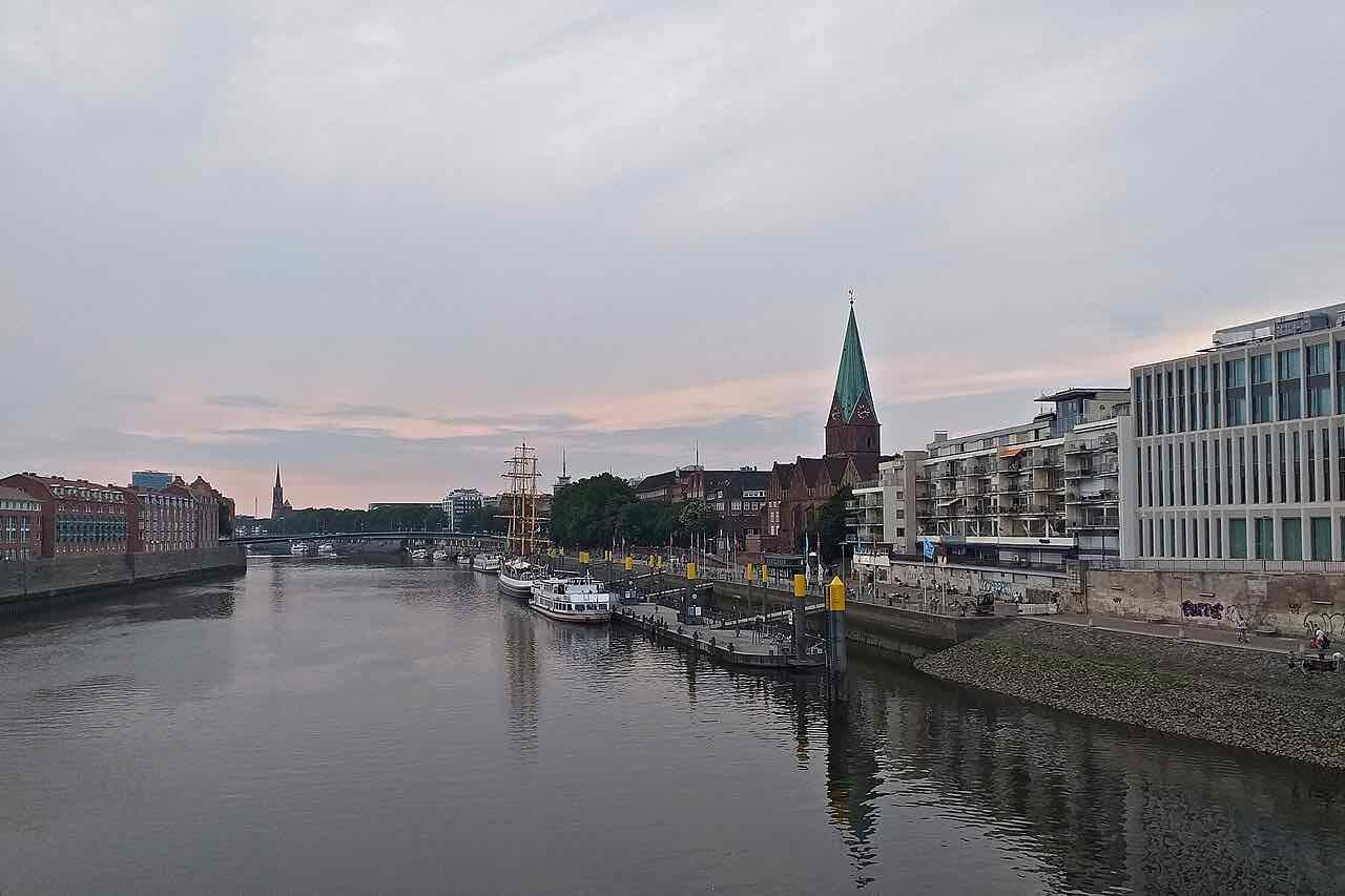 The Weser at dusk seen from a bridge, with moored boats and a tall-masted ship along the embankment and a green church spire on the right bank