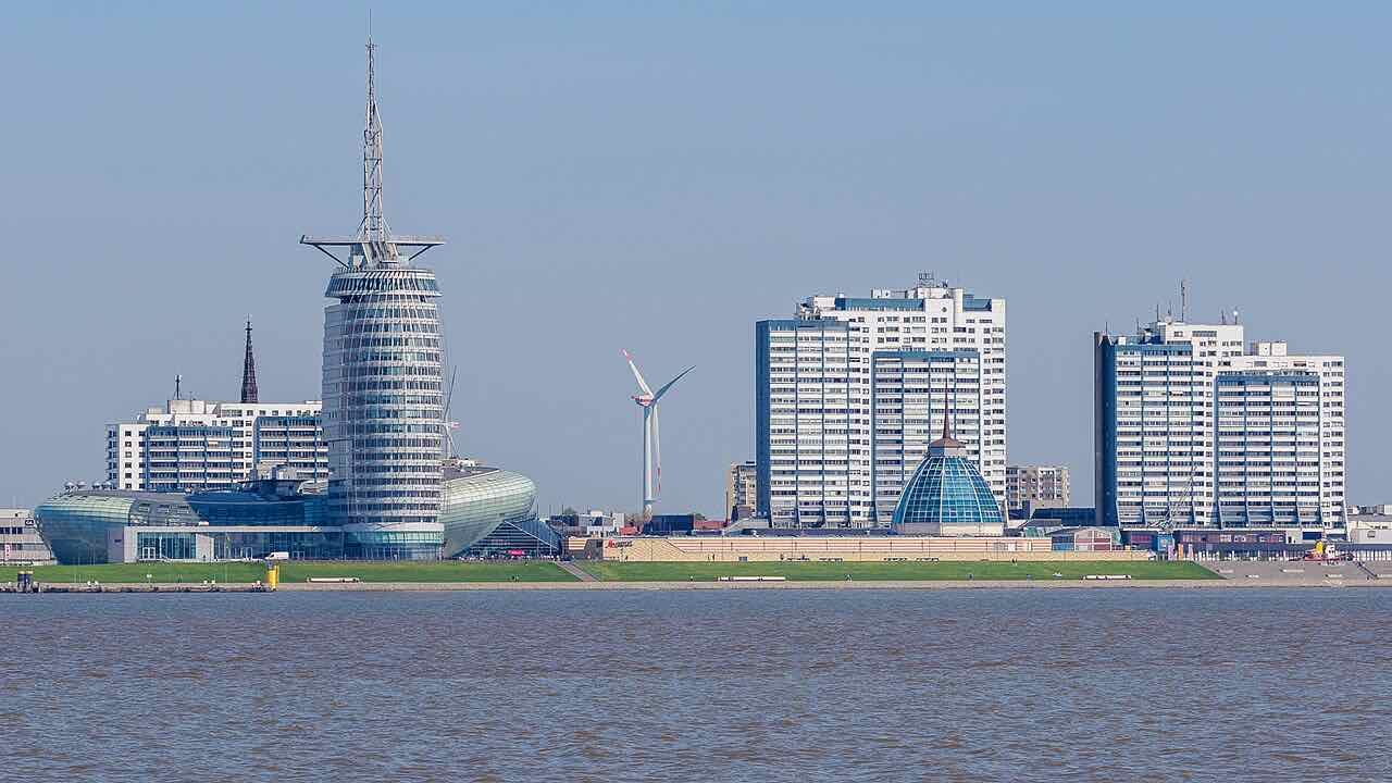 Bremerhaven's waterfront seen across the brown water of the Weser: the slim round tower of the Atlantic Hotel Sail City, the curved glass roof of the Klimahaus, a glass dome, a wind turbine and white high-rise blocks behind a grass-covered dyke