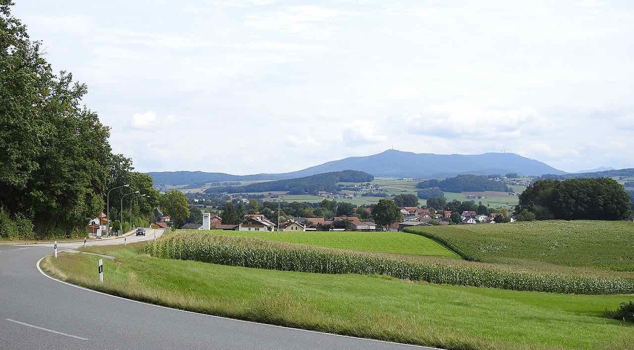 A country road curving past a maize field towards the village of Kothmaißling, with the long ridge of the Hoher Bogen on the horizon