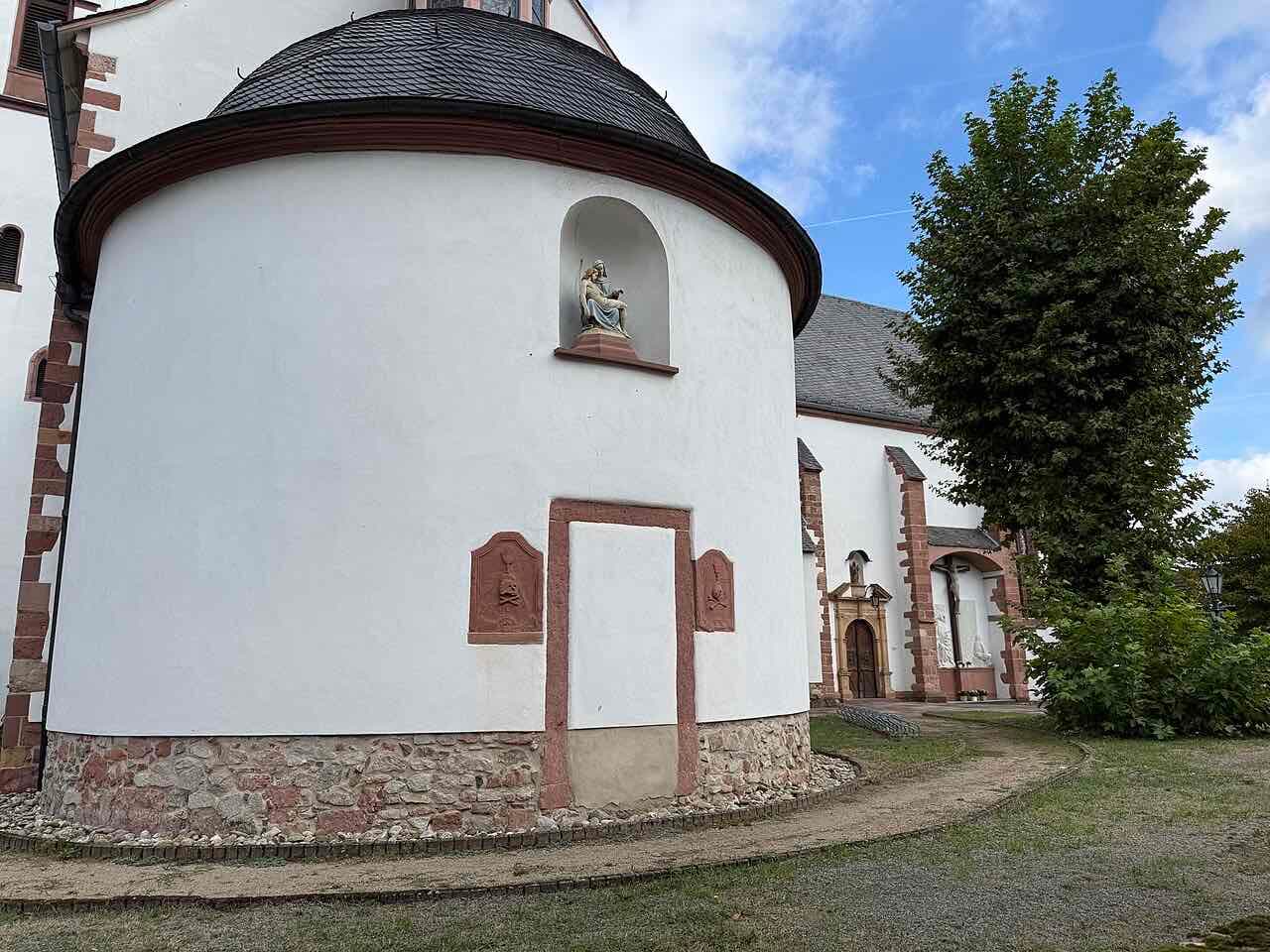 The white rounded apse of the Wallfahrtskirche in Dieburg with a red sandstone door frame, a small Pietà figure in a niche and a lime tree beside it