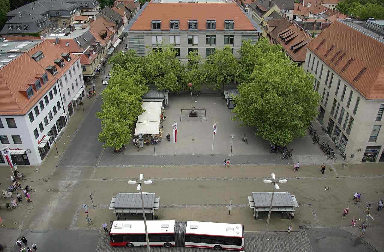 The Hugenottenplatz in Erlangen from above, a paved square with plane trees, a café awning and bus stops in front