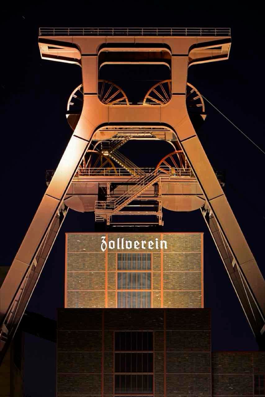 The Zollverein shaft 12 winding tower lit up at night with the word Zollverein on the building below