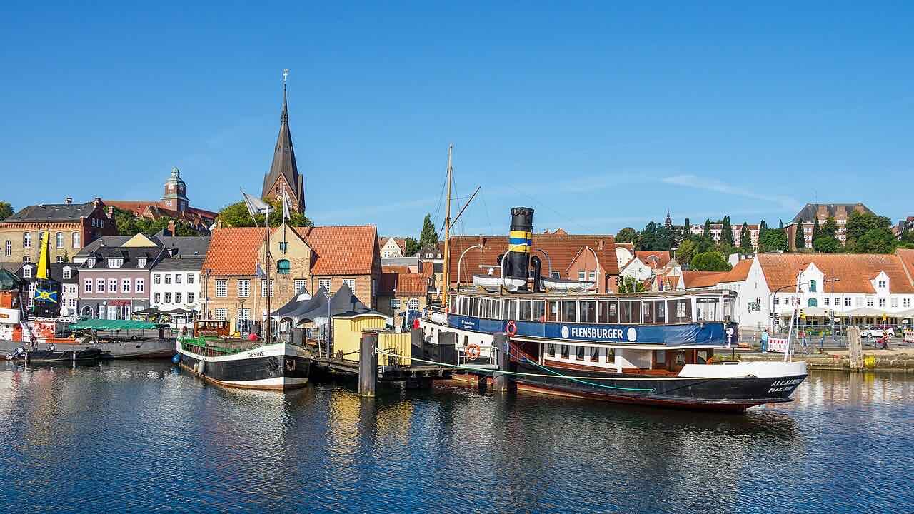 The historic steamer Alexandra and a sailing barge at the Schiffbrücke quay in Flensburg harbour, with red-roofed houses and the spire of St. Marien behind them under a blue sky