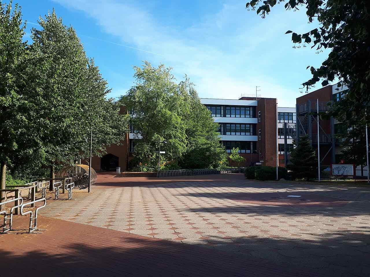 A paved courtyard with bicycle racks in front of red-brick and white teaching buildings of Hochschule Flensburg, framed by trees