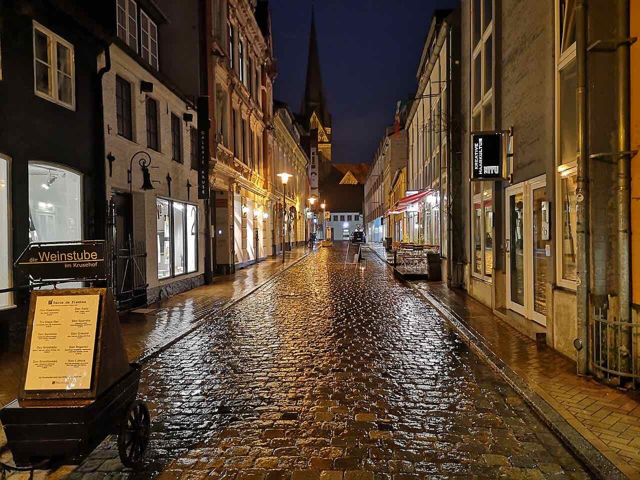 A wet cobbled street in the Flensburg old town at night, lit by street lamps and shop windows, with a church spire at the far end