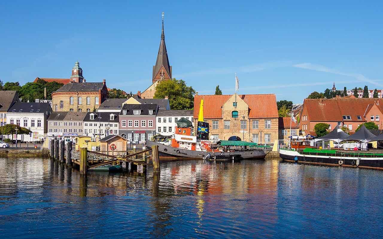 Old harbour buildings, a tugboat and a landing stage on the water at the Schiffbrücke, with the tall dark spire of St. Marien rising behind the houses