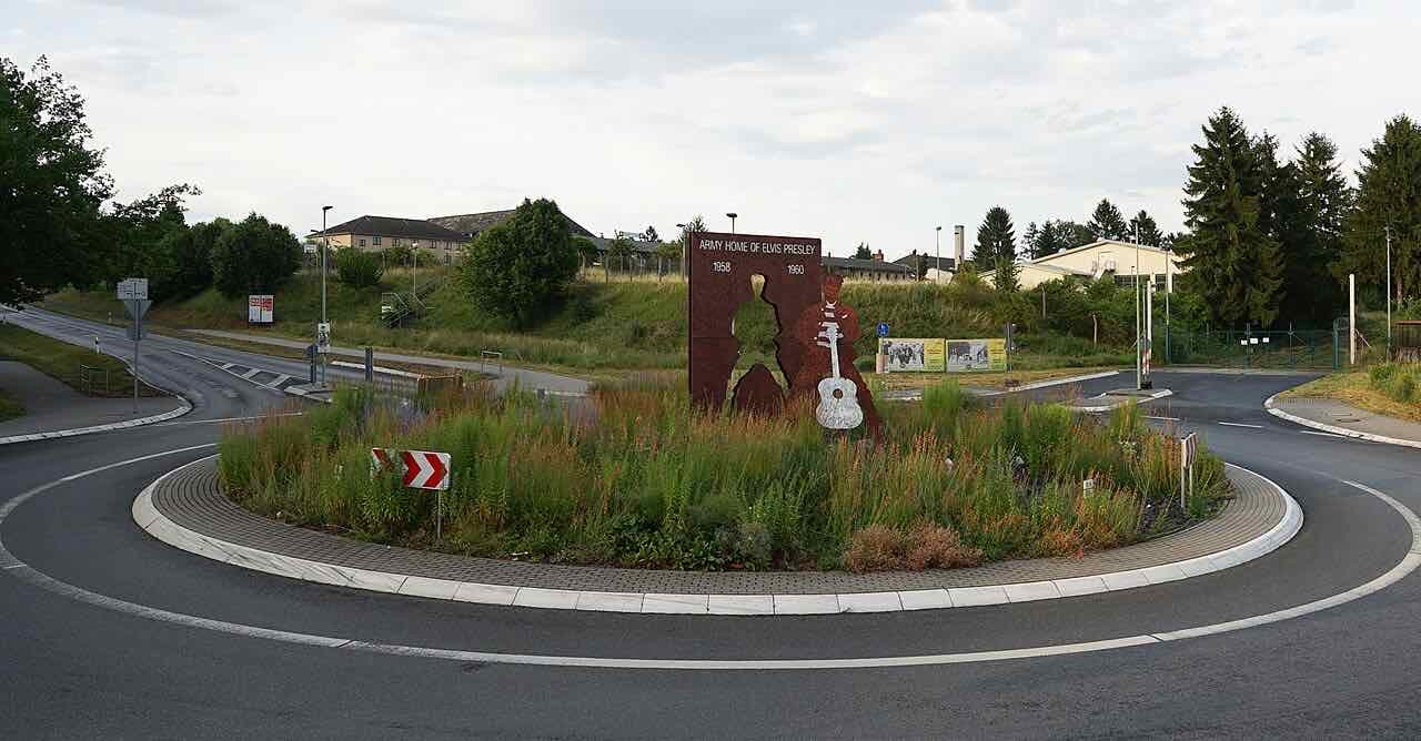A rust-coloured steel monument with a guitar and the words Army Home of Elvis Presley 1958 1960 on a roundabout planted with tall grasses