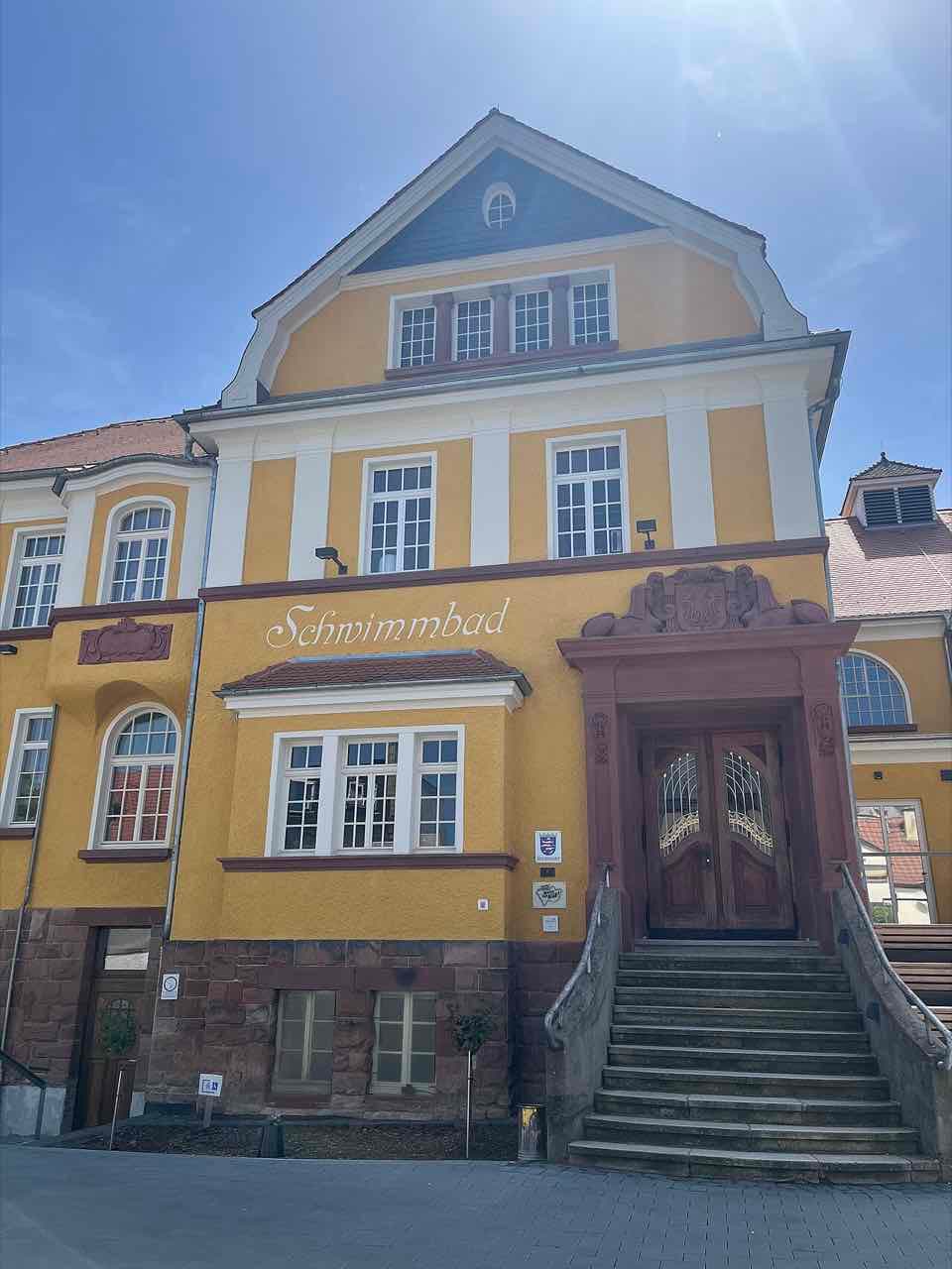 The ochre front of the former indoor swimming pool in Friedberg with the word Schwimmbad on the facade and a carved wooden double door at the top of a stone staircase
