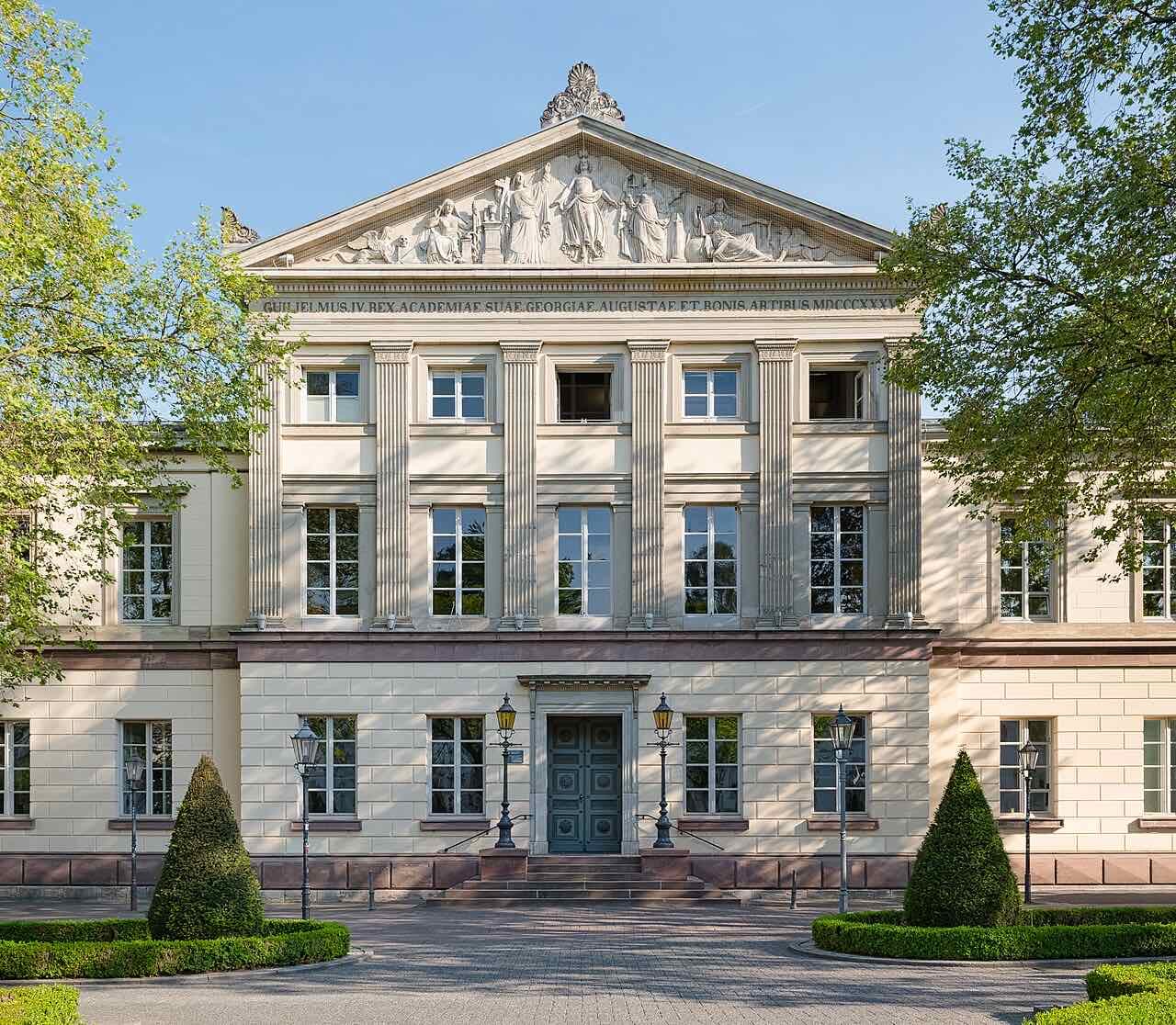 The pale classical front of the university Aula on Wilhelmsplatz, with a carved triangular pediment, columns and clipped box trees in front