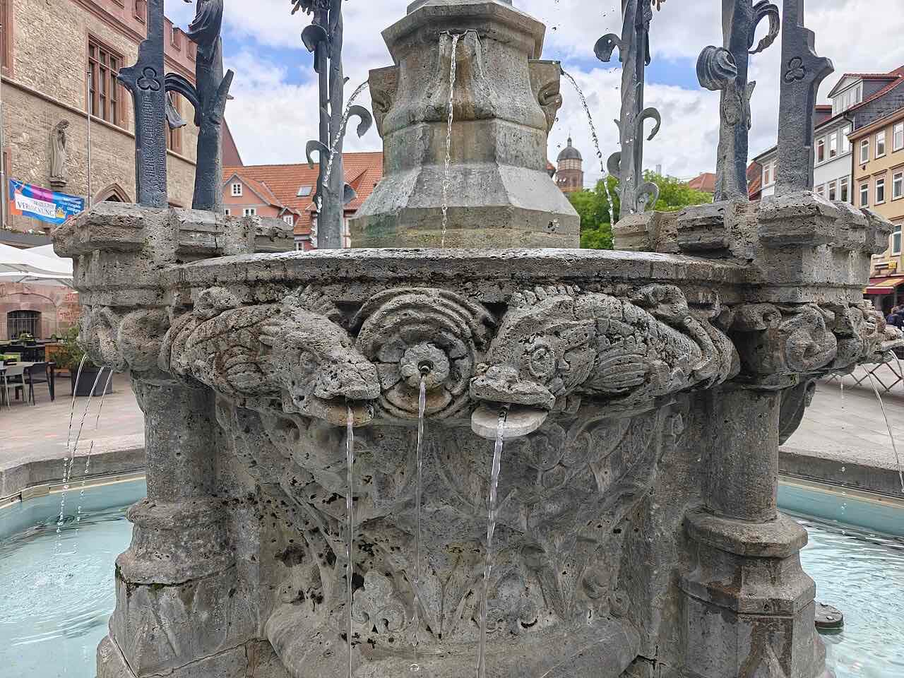 The carved stone basin of the Gänseliesel fountain with water running from three spouts, and the iron canopy above it