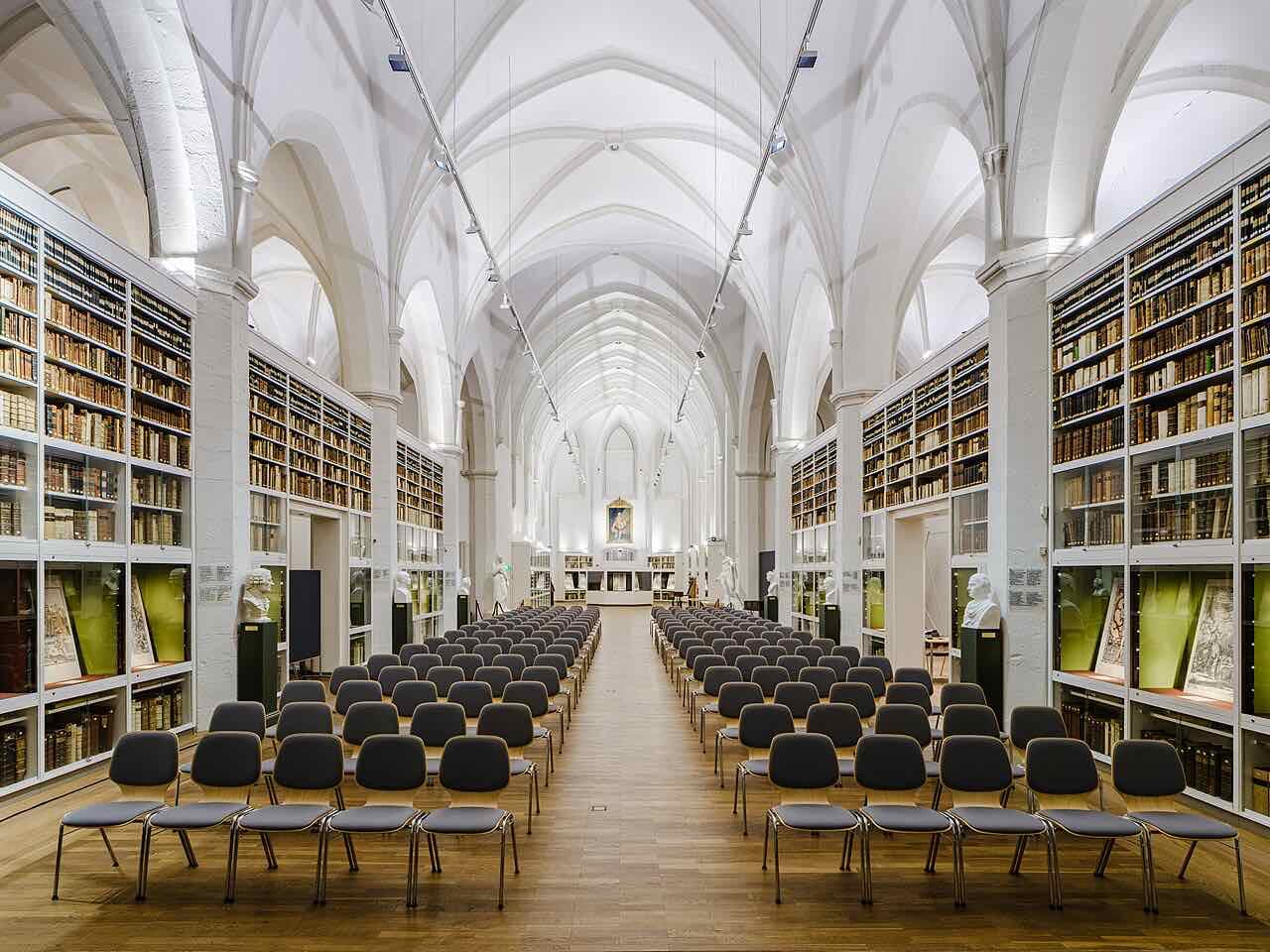 The white vaulted interior of the Paulinerkirche in Göttingen with glass-fronted shelves of old books on both sides and rows of chairs in the nave