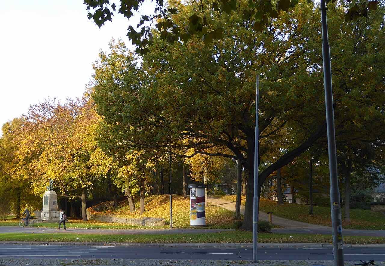 The grassy Stadtwall rampart at the Groner Tor under autumn trees, with a small monument, a cyclist and a walker beside the road