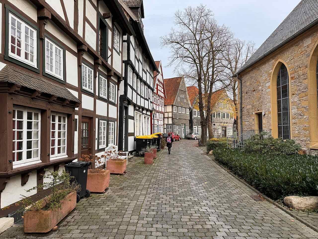 A cobbled lane in Gütersloh's old village core: half-timbered houses with white-framed windows on the left, a sandstone church wall with a tall pointed window on the right, and bare trees ahead