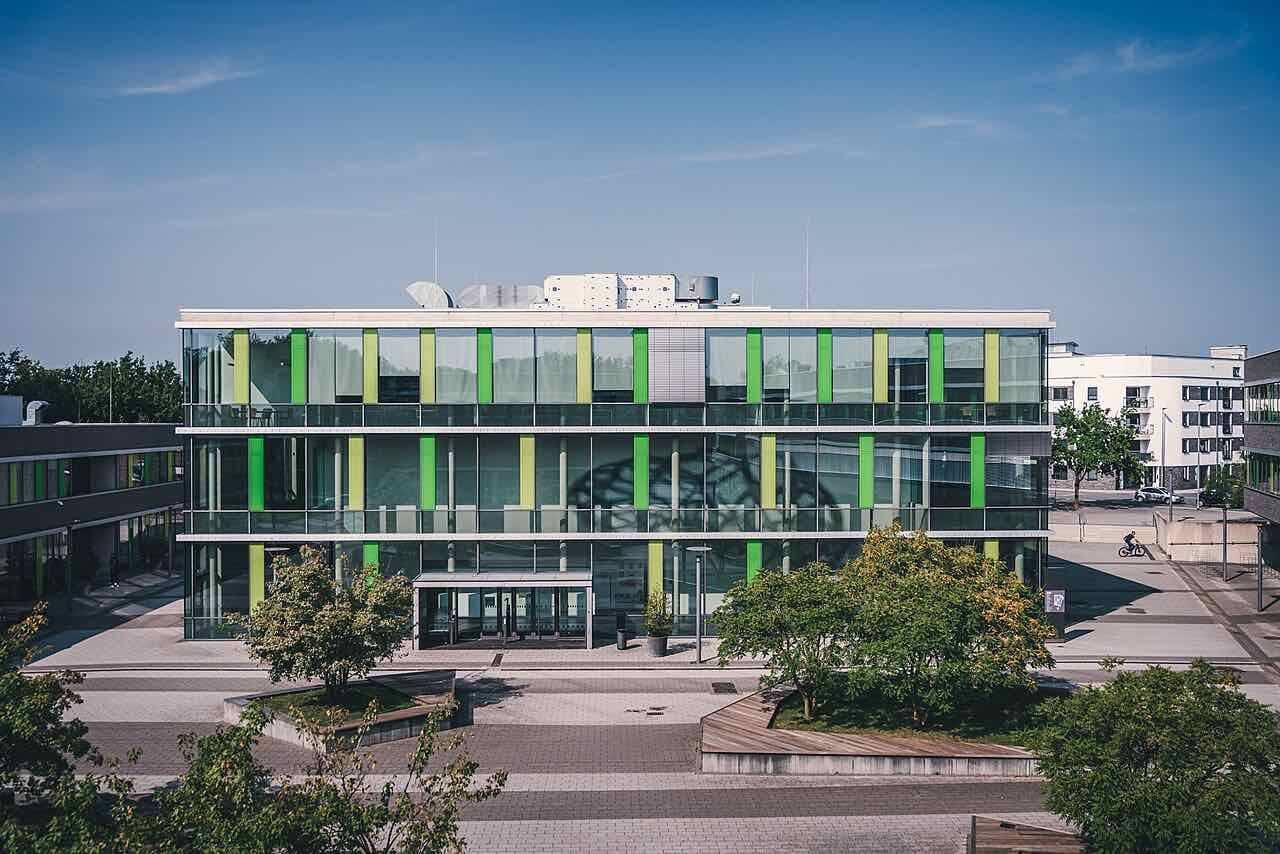 A modern glass university building with green and yellow facade strips on the Kamp-Lintfort campus, with trees and a paved square in front