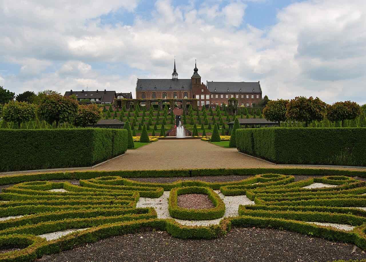 Kloster Kamp seen over the low box-hedge ornaments of the baroque garden, with a fountain on the central axis and a partly cloudy sky