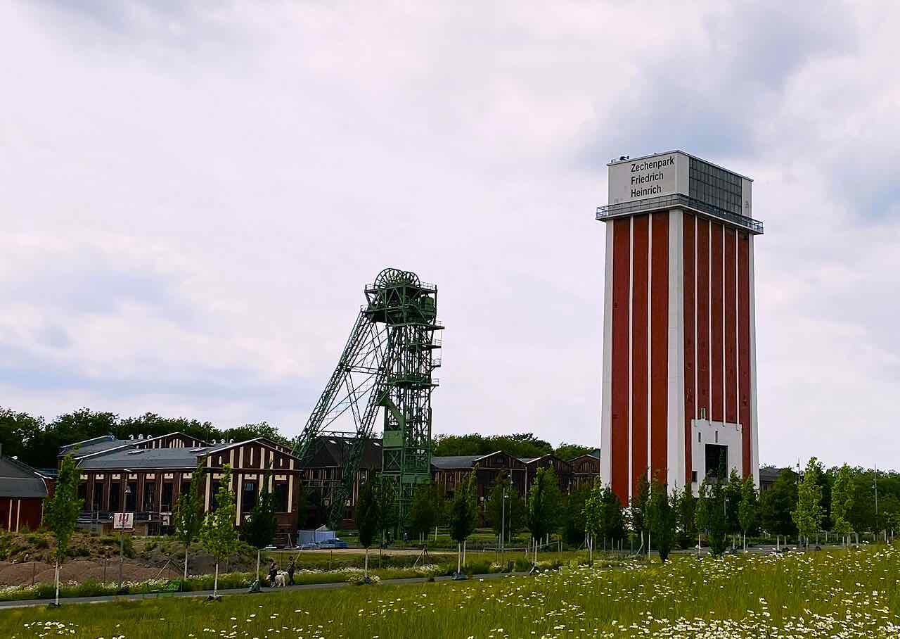 The Zechenpark Friedrich Heinrich: old colliery halls, a green headframe and the red and white tower under a cloudy sky, with a meadow and walkers in front