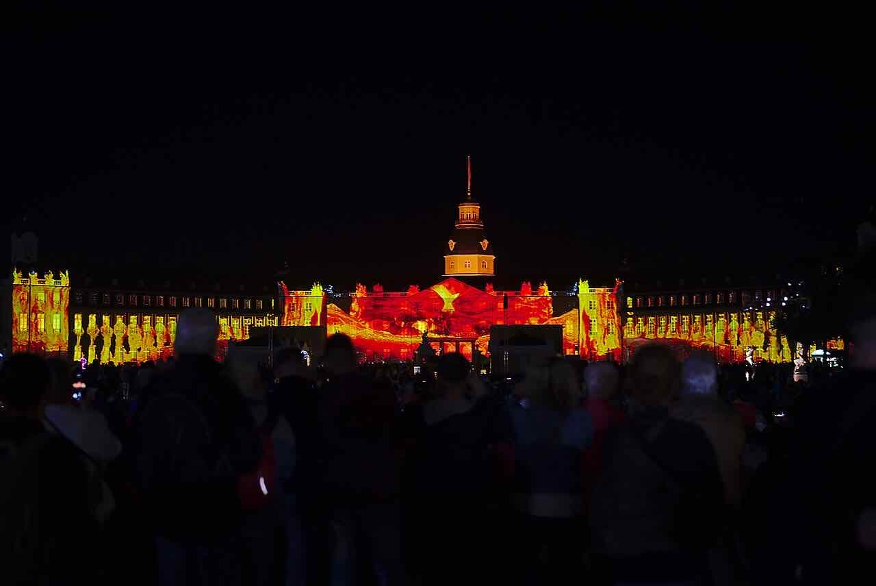 The palace facade covered in red and orange light projections at night, with a crowd in dark silhouette in front