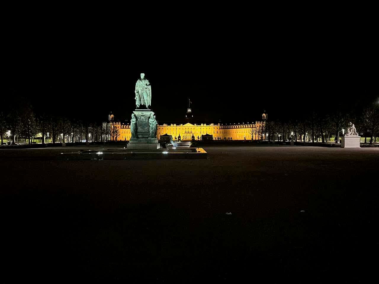 Karlsruhe Palace lit yellow at night behind a statue on the empty Schlossplatz