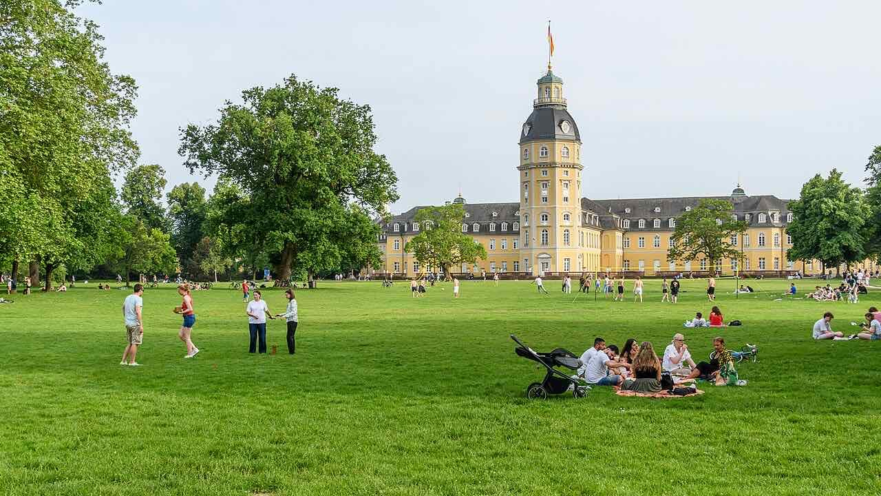 People sitting and walking on a large lawn in the Karlsruhe palace garden, with the palace tower behind big trees