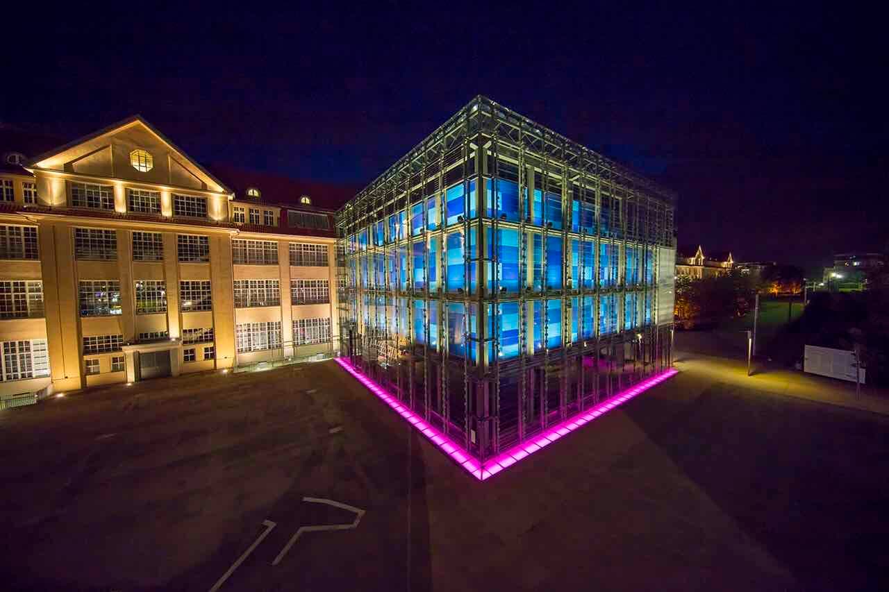The glass cube of the ZKM lit blue and pink at night next to an old brick factory building