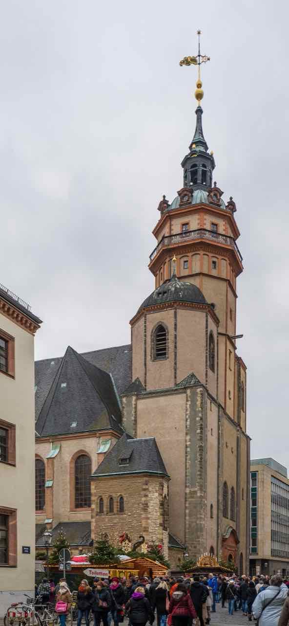 The tower of the Nikolaikirche in Leipzig under a grey sky, with a crowd and market stalls at its foot