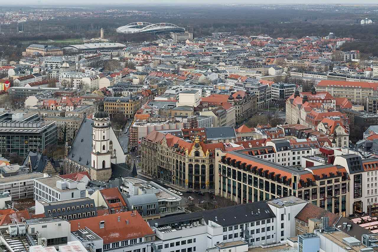 The Leipzig city centre from above, with the white tower of the Thomaskirche among red roofs and a stadium and green space behind the houses