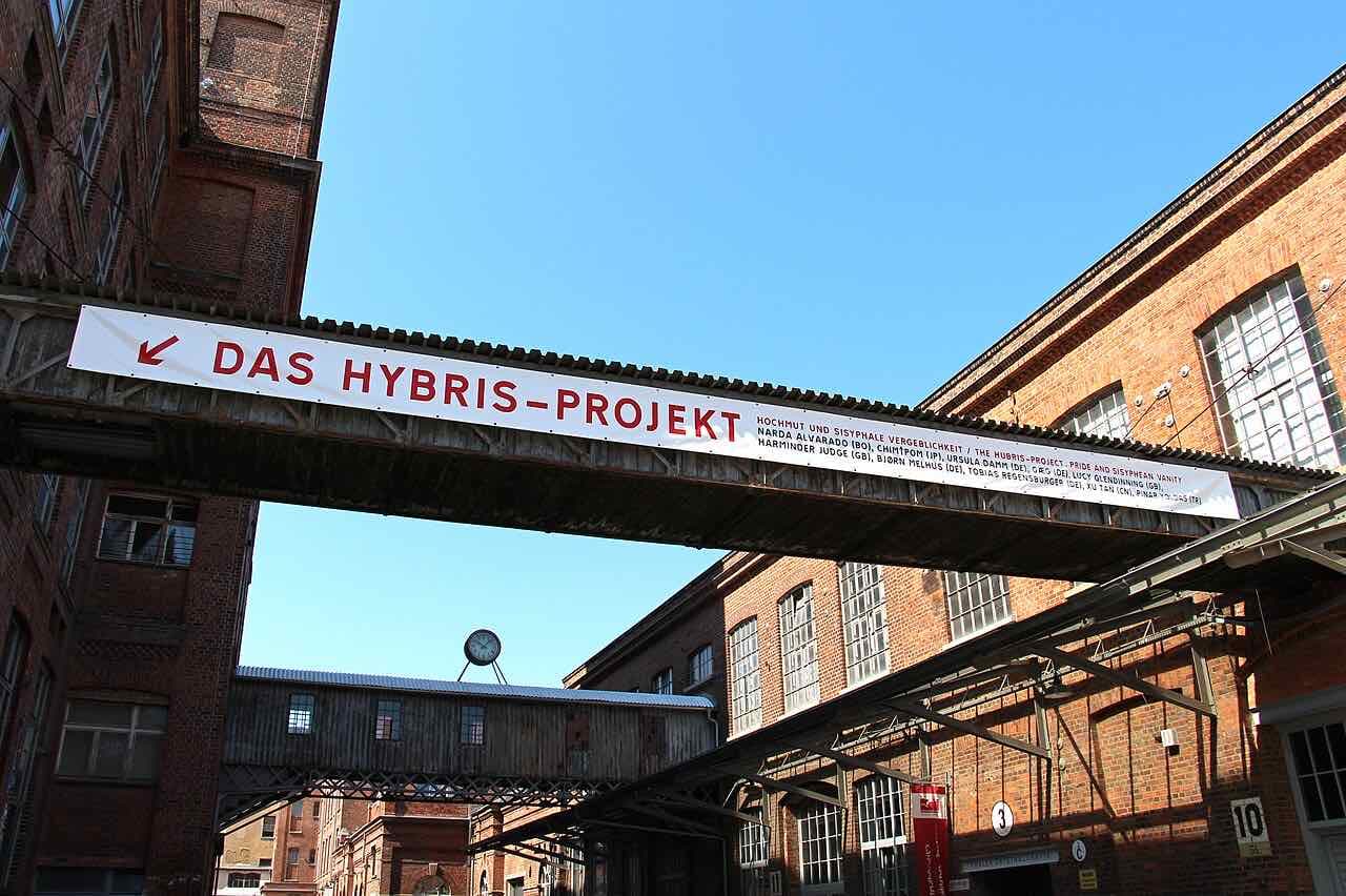 Red-brick factory buildings of the Baumwollspinnerei joined by a covered walkway under a blue sky, with an exhibition banner on it