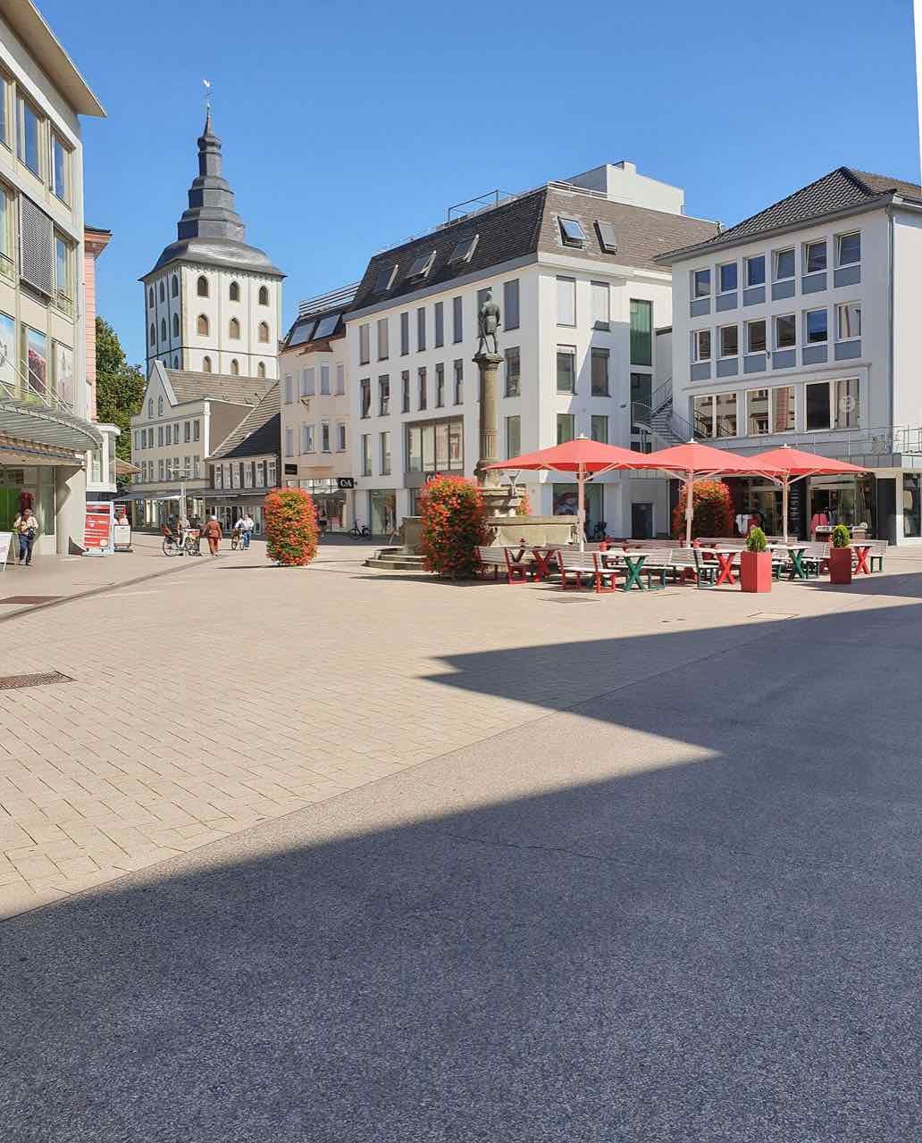 The pedestrian zone on Lange Straße in Lippstadt on a sunny day, with the Bernhard fountain, red café umbrellas and the grey tower of the Jakobikirche behind the houses