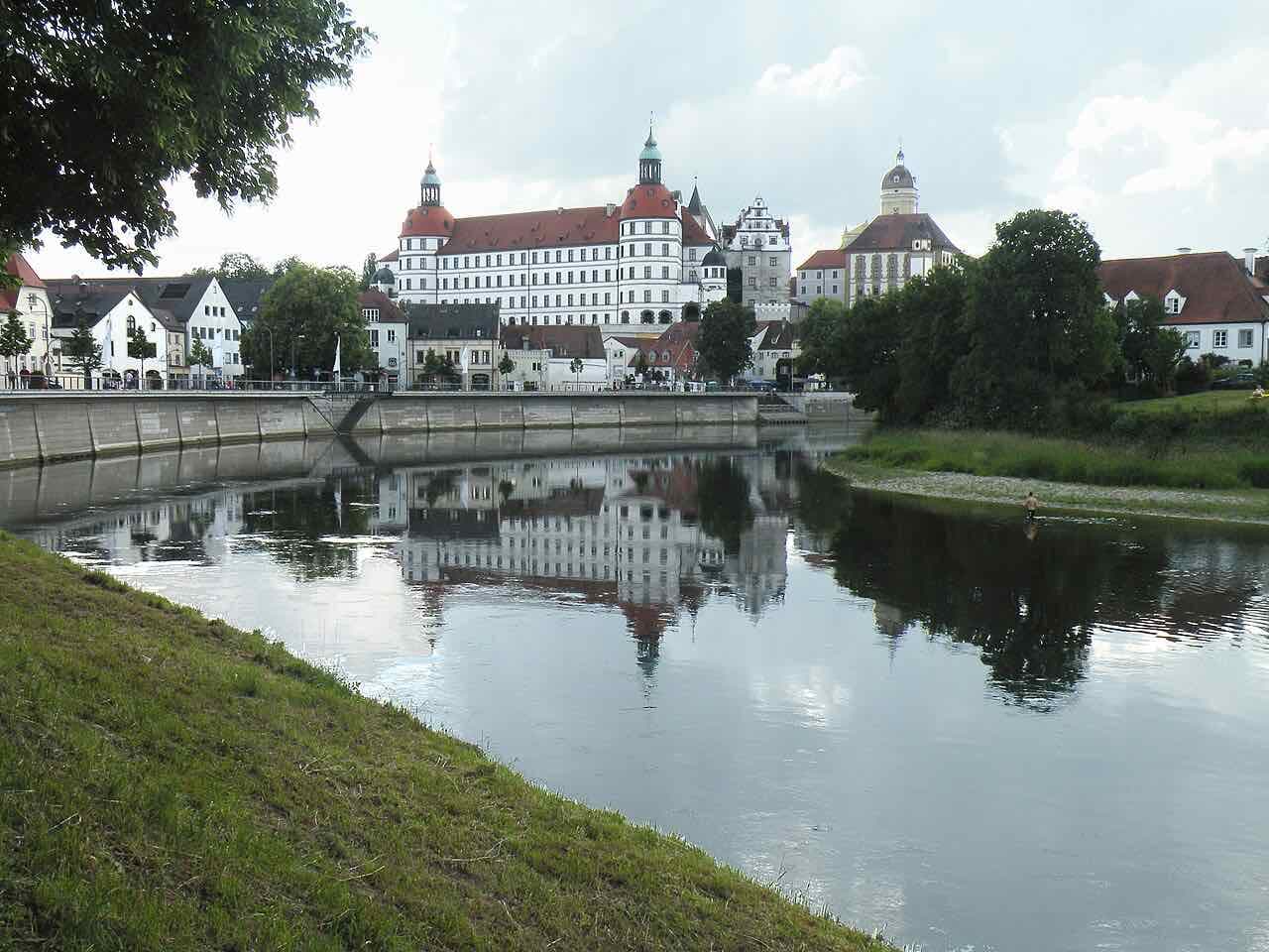 The white Renaissance palace of Neuburg with red roofs and green-capped towers, standing above the old town and reflected in the calm Danube in the foreground
