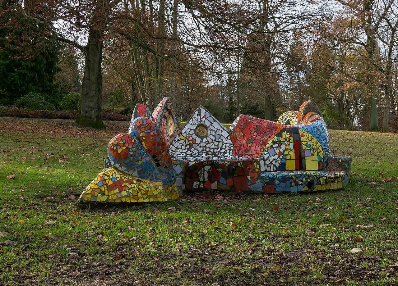 A bench covered in colourful mosaic tiles on a lawn under bare autumn trees in the Bürgerpark