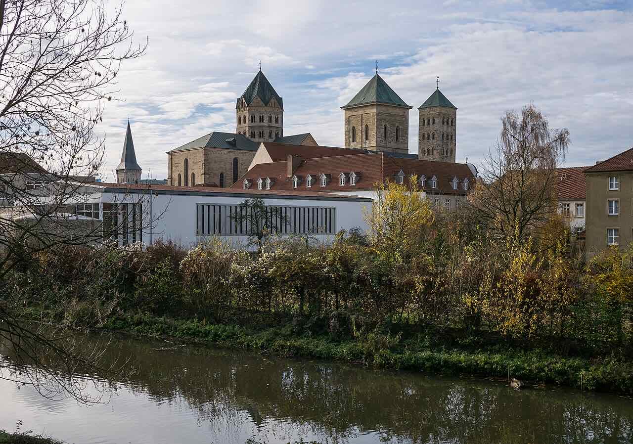 The river Hase running quietly through the old town in autumn, with the stone towers of a Romanesque church rising behind the trees