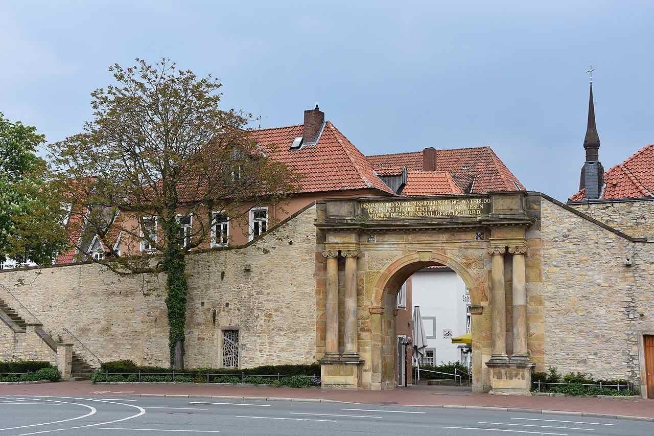 The Heger Tor, a sandstone gate with columns and a gold inscription set into a stretch of old city wall, with red-tiled roofs behind it
