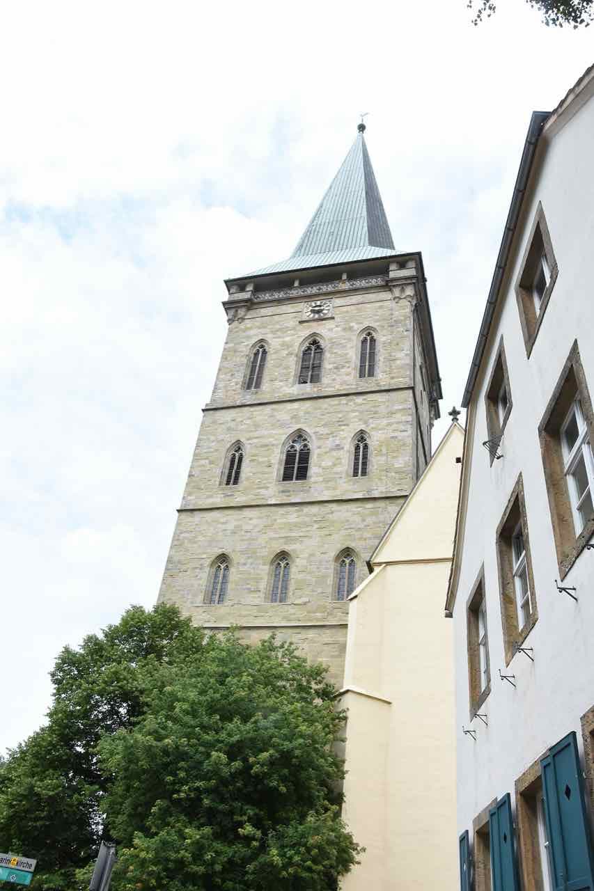 The tall stone tower of St Katharinen church with a green spire, seen from a narrow street between old houses