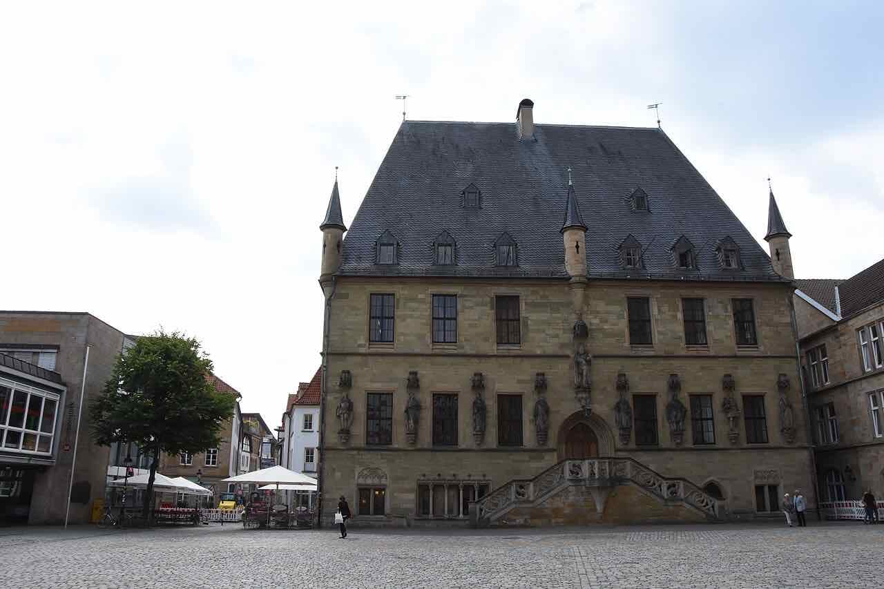 The stone town hall of Osnabrück on its cobbled market square, with a steep slate roof, three slim corner turrets, a row of statues on the facade and an outside staircase