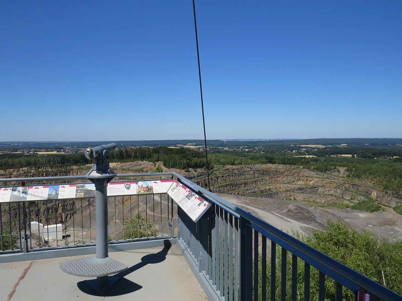 A viewing platform with a coin telescope and information boards looking over the terraced rock walls of the Piesberg quarry and the flat countryside beyond
