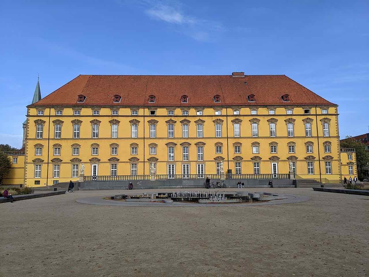 The long yellow Baroque palace of Osnabrück with a red roof and many tall windows, seen across a gravel square with a low fountain