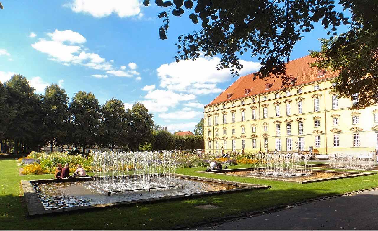 Flower beds and two rectangular fountain pools in the palace garden on a sunny day, with the yellow palace wing on the right and people sitting on the grass