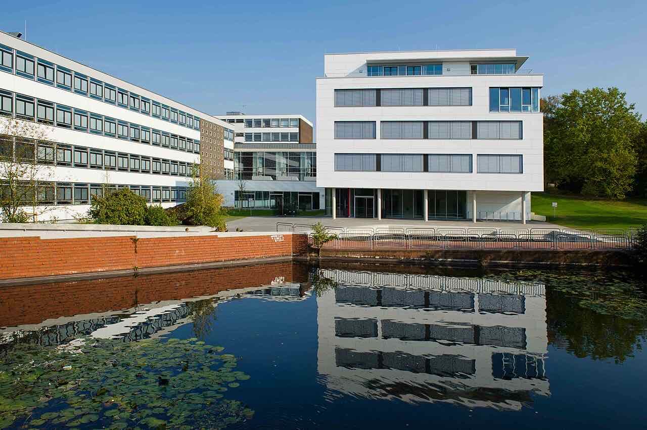 White and glass faculty buildings of the Hochschule Osnabrück on the Westerberg campus, reflected in a lily pond in front of a brick wall