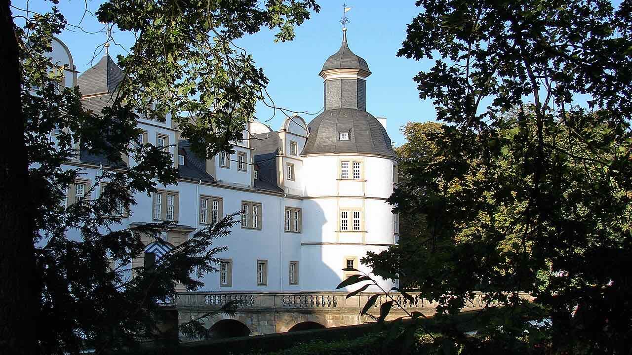 The white round tower and façade of Schloss Neuhaus seen through trees, with a stone bridge in front