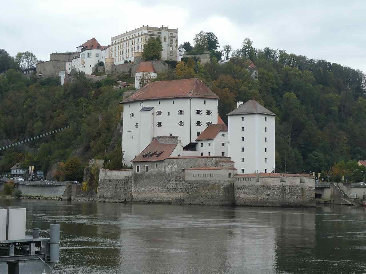 The stone Niederhaus on the waterfront under a cloudy sky, with the long pale Oberhaus fortress on the wooded hill behind