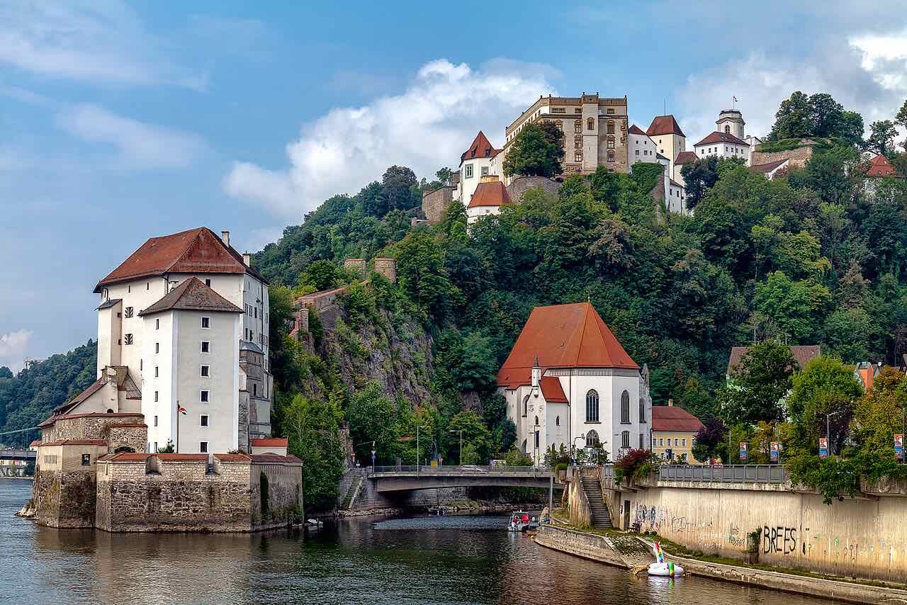 The white walls of Veste Niederhaus beside the river with a stone bridge, a small church and the Oberhaus fortress on the wooded hill above