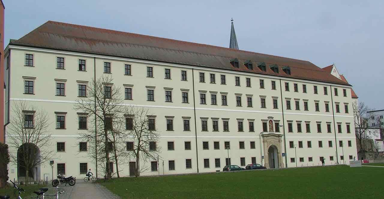The long cream-coloured Nikolakloster building with a red roof and a church spire behind it, on a lawn in early spring