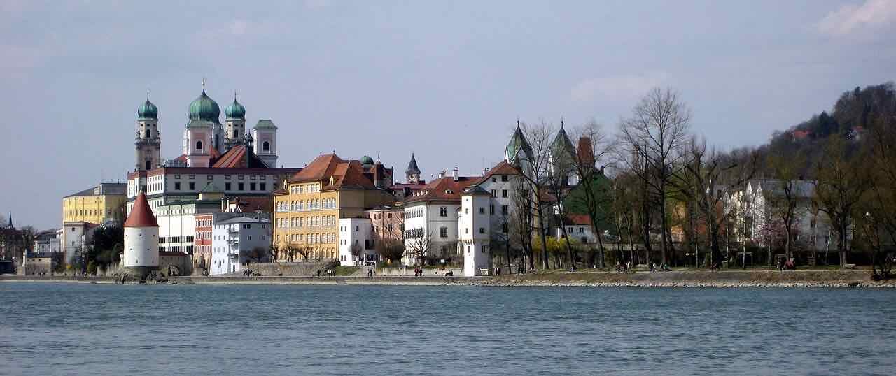 The old town of Passau seen across the water, with the green domes of the cathedral, a round white tower and a row of coloured houses along the bank
