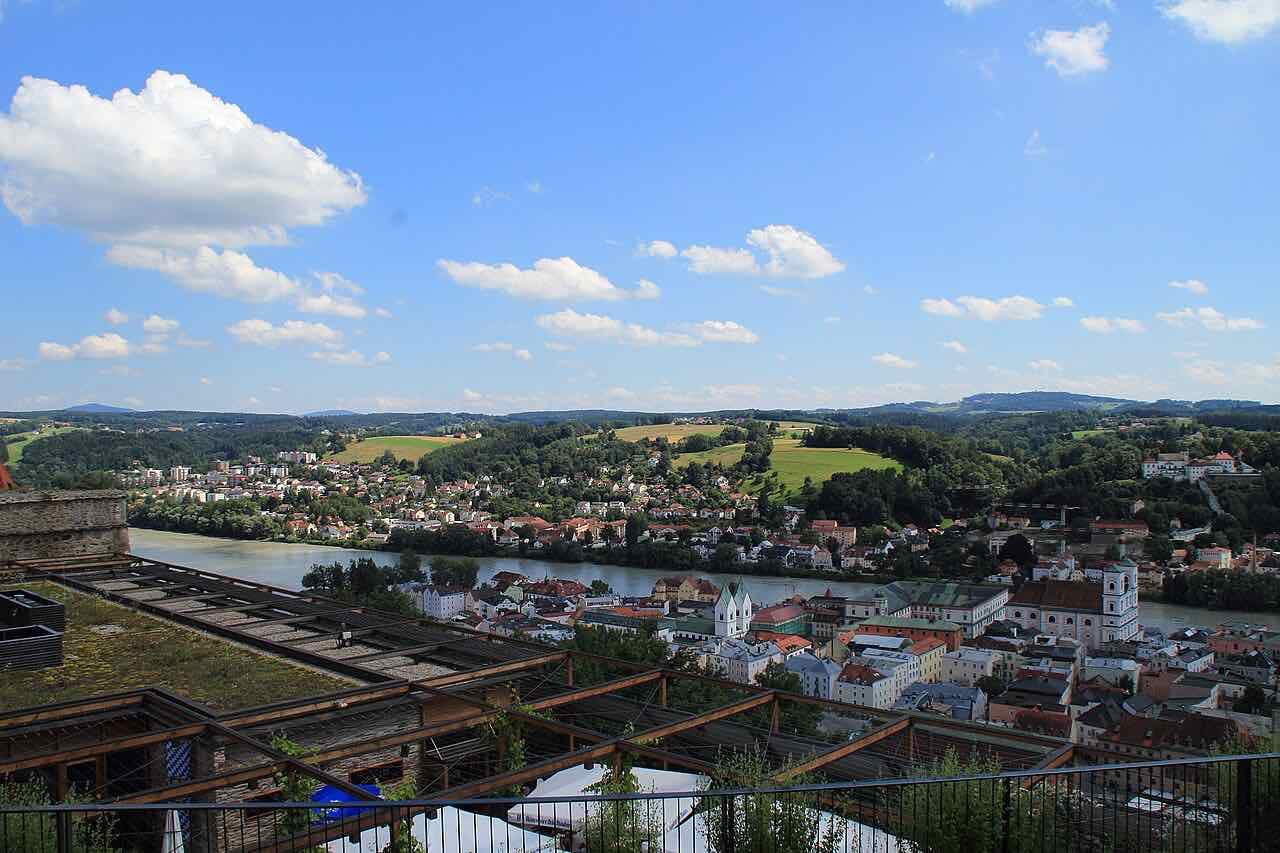 A view over the roofs of Passau and a wide green river valley under a blue sky with white clouds, seen from a high terrace