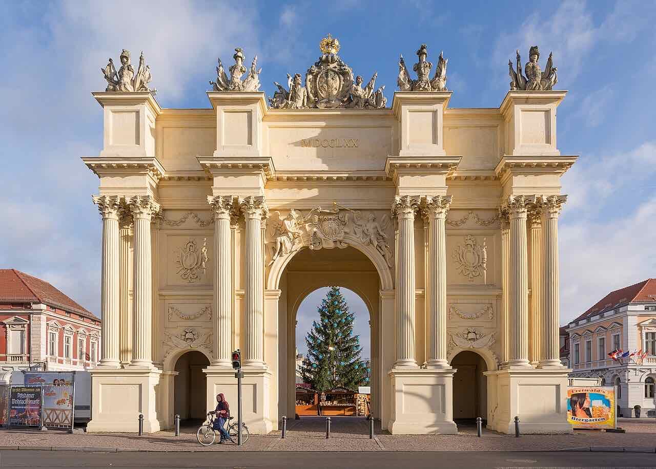 The Brandenburg Gate in Potsdam, a cream-coloured triumphal arch with columns and sculptures, with a Christmas tree behind it and a cyclist in front