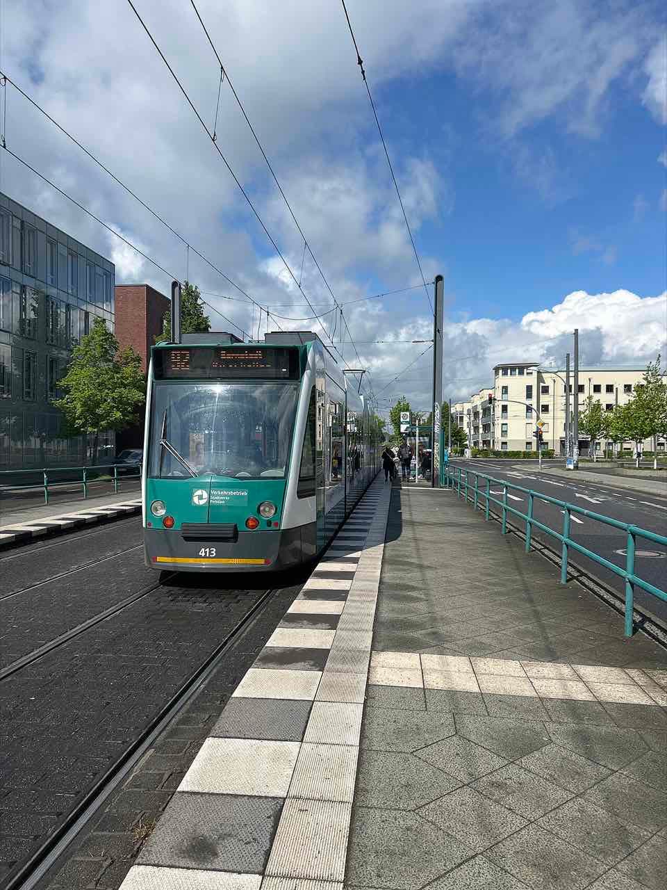 A green and white Potsdam tram at a stop beside a glass-fronted building on the Fachhochschule campus
