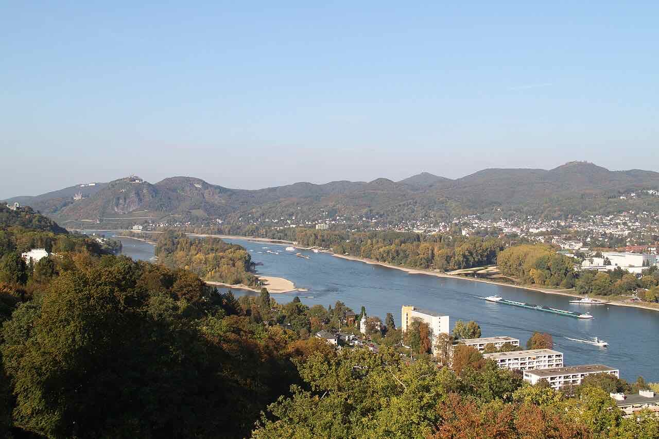 The Rhine from a lookout above Rolandseck: the river bends past a wooded island with a sandbank, a cargo barge and a ferry on the water, and the hills of the Siebengebirge with Drachenfels castle rise on the far bank