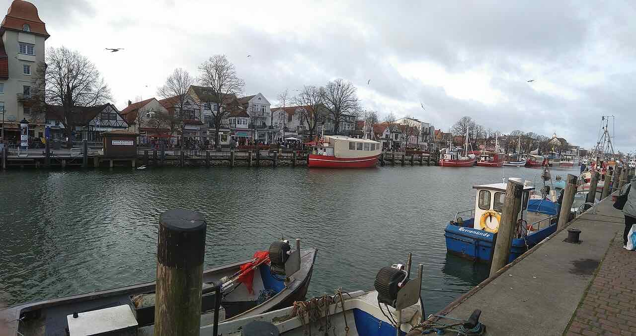 Fishing boats and small ferries moored along the Alter Strom channel in Warnemünde, with a row of cafés and half-timbered houses on the far bank