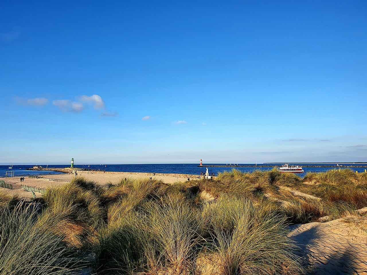 Dune grass in the foreground and a sandy beach beyond, with the mouth of the Warnow and two small lighthouses under a deep blue sky